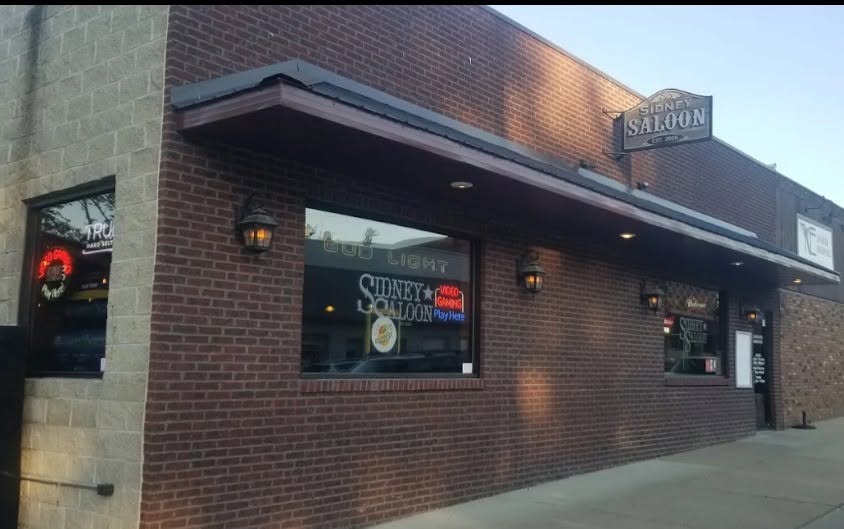 The brick storefront of the Sidney Saloon at dusk, with its hanging sign and lit lanterns